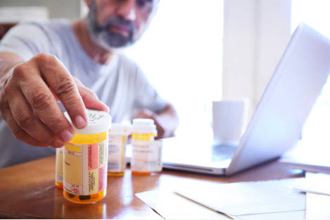 Senior man looking at a pill bottle while working on a laptop